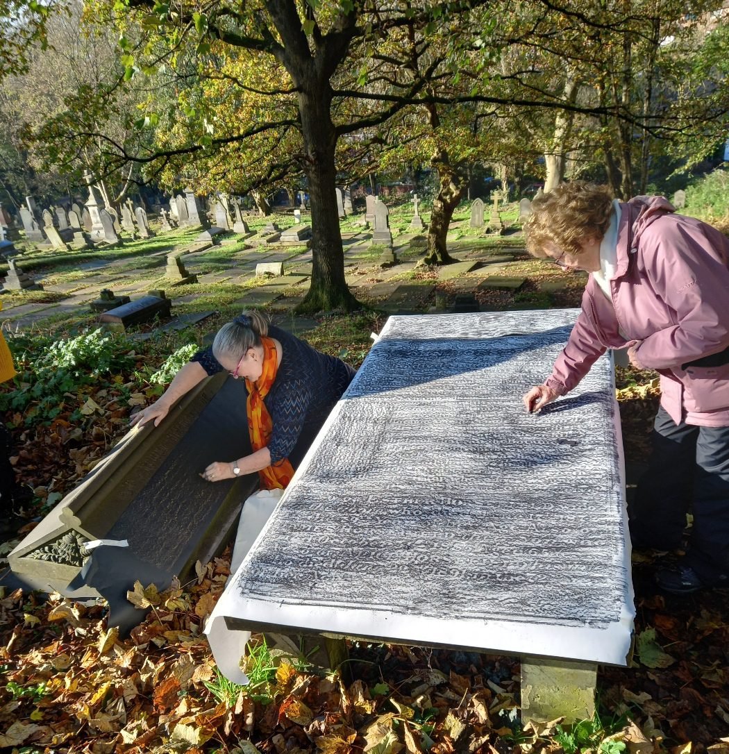 Two women creating gravestone rubbings at a table in St Mary’s Churchyard, Prestwich, with rows of headstones behind them