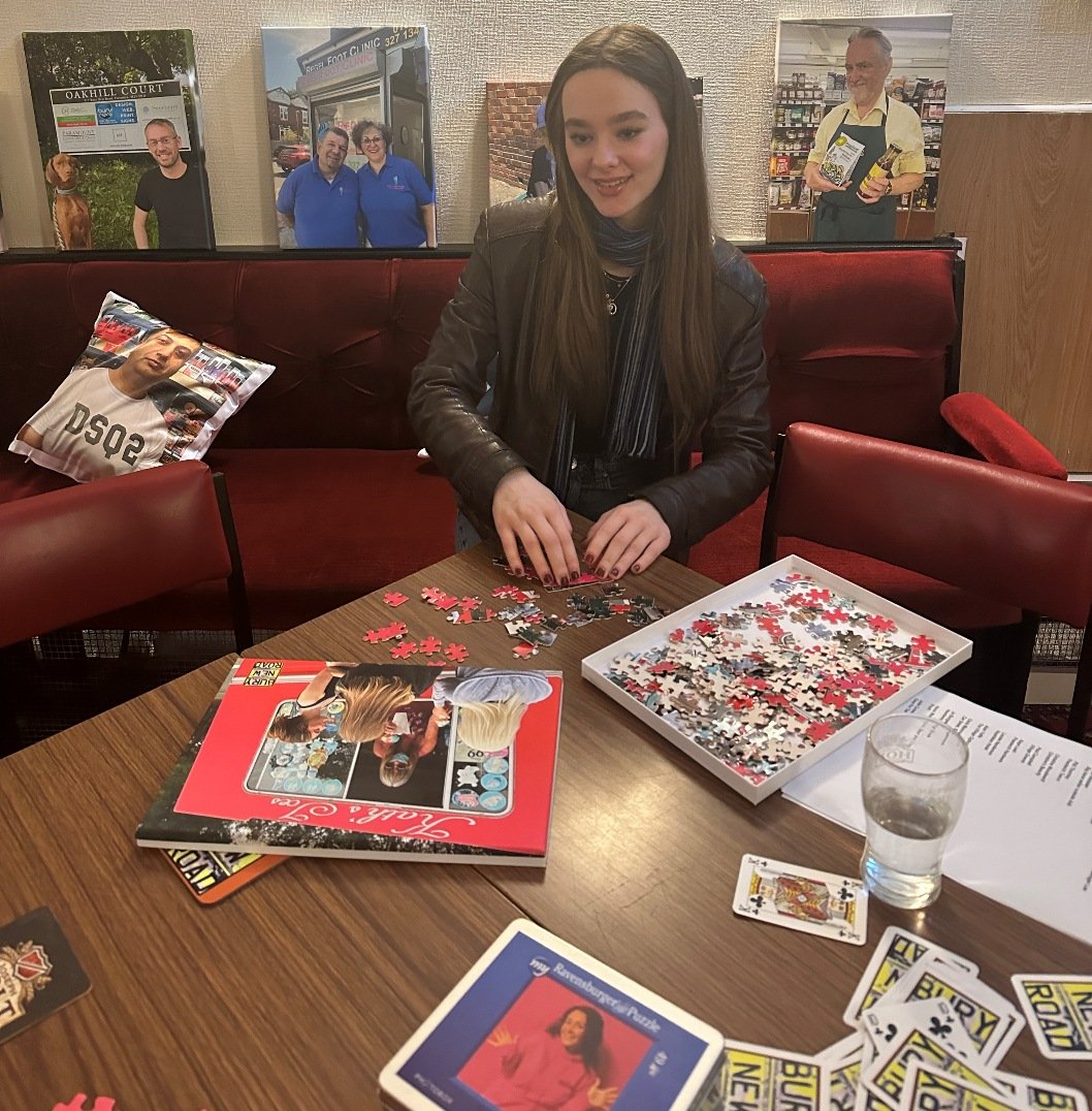 Demi piecing together a Portraits Exhibition jigsaw, with the portrait playing cards and a cushion printed with a portrait on the seat beside her