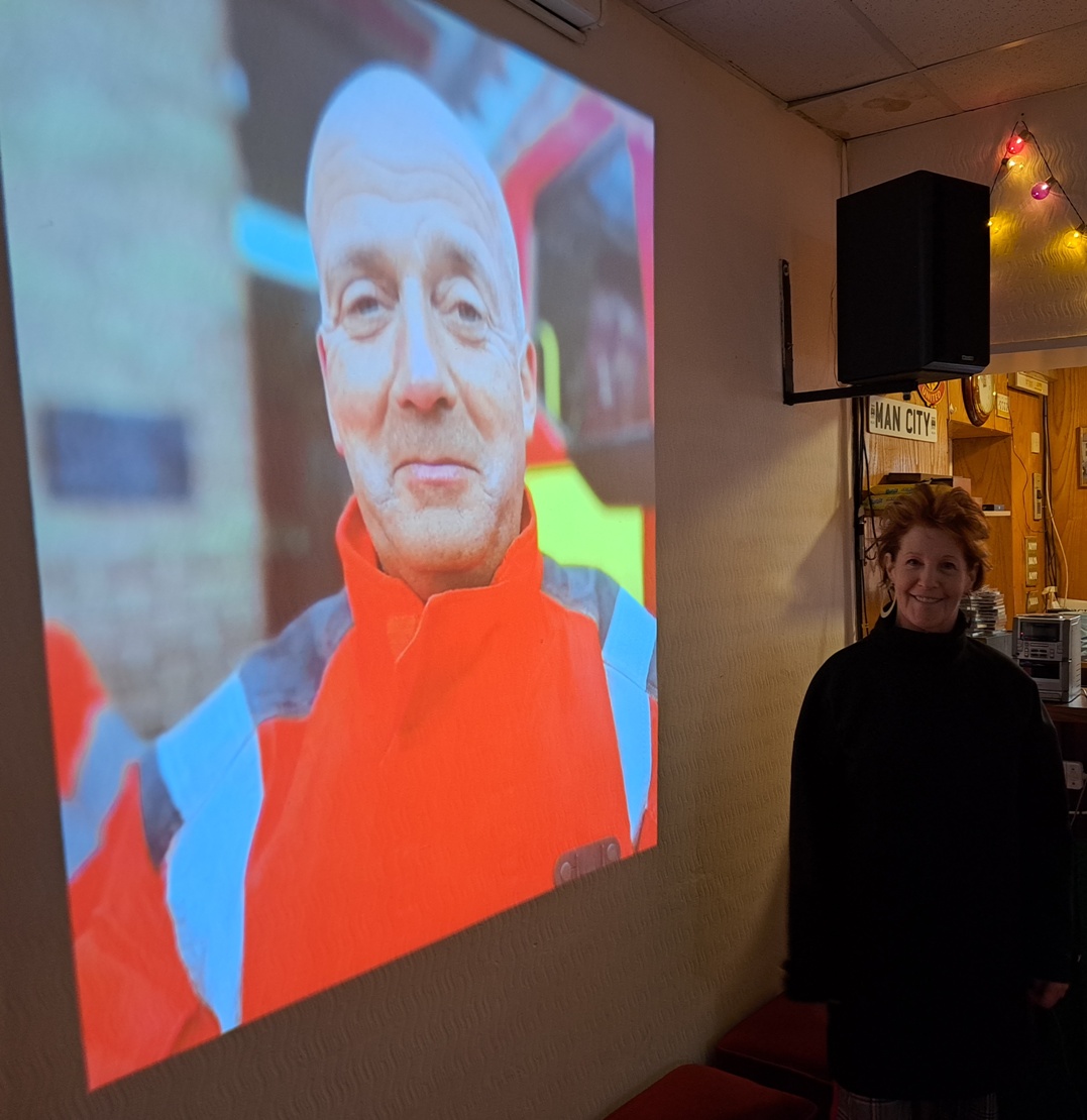 A portrait from the Portraits Exhibition projected on the wall &ndash; a man in an orange and blue hi-vis jacket &ndash; with Louise Garman standing beside it