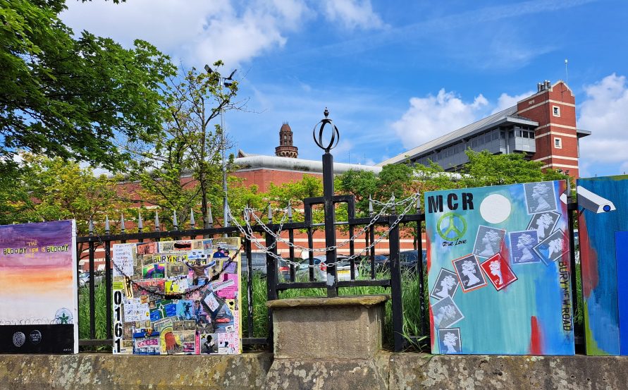 Four canvases chained to the railings: a purple sunset skyline, a mixed-media collage board, an &lsquo;MCR One Love&rsquo; postage-stamp piece and a blue abstract painting