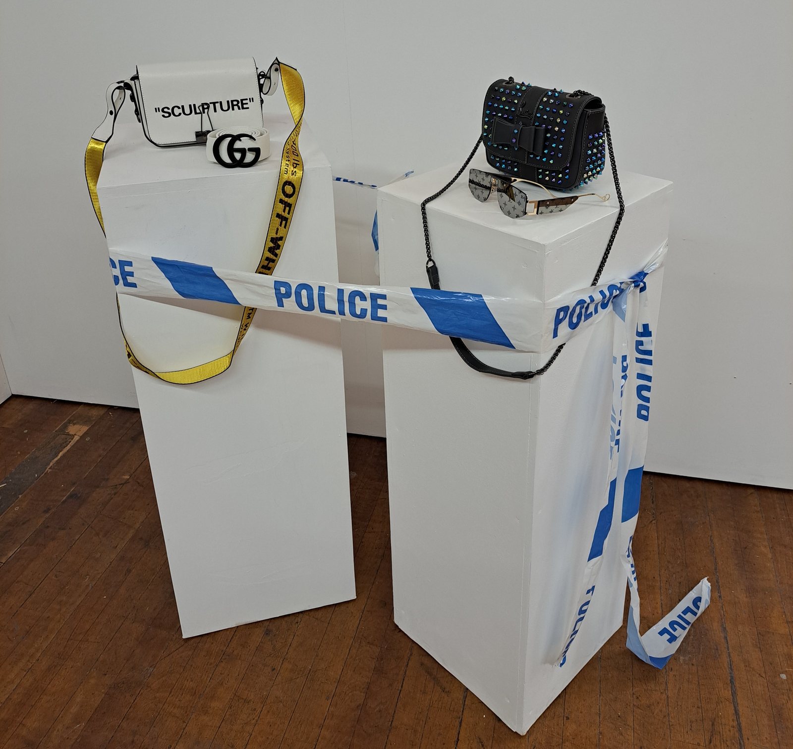 The &lsquo;Counterfeit Street&rsquo; installation: two gallery plinths displaying a fake Off-White bag marked &lsquo;Sculpture&rsquo;, a Gucci belt, a studded bag and sunglasses, wrapped in police tape