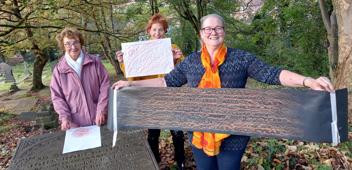 Three women in a Prestwich churchyard holding up a headstone rubbing during a gravestone-rubbing workshop