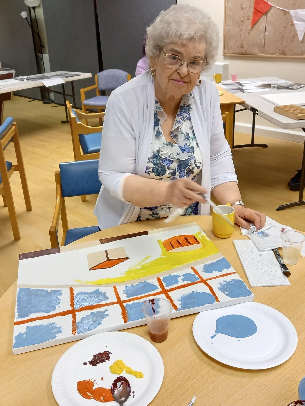 An elderly woman painting a stylised townscape canvas at a community workshop