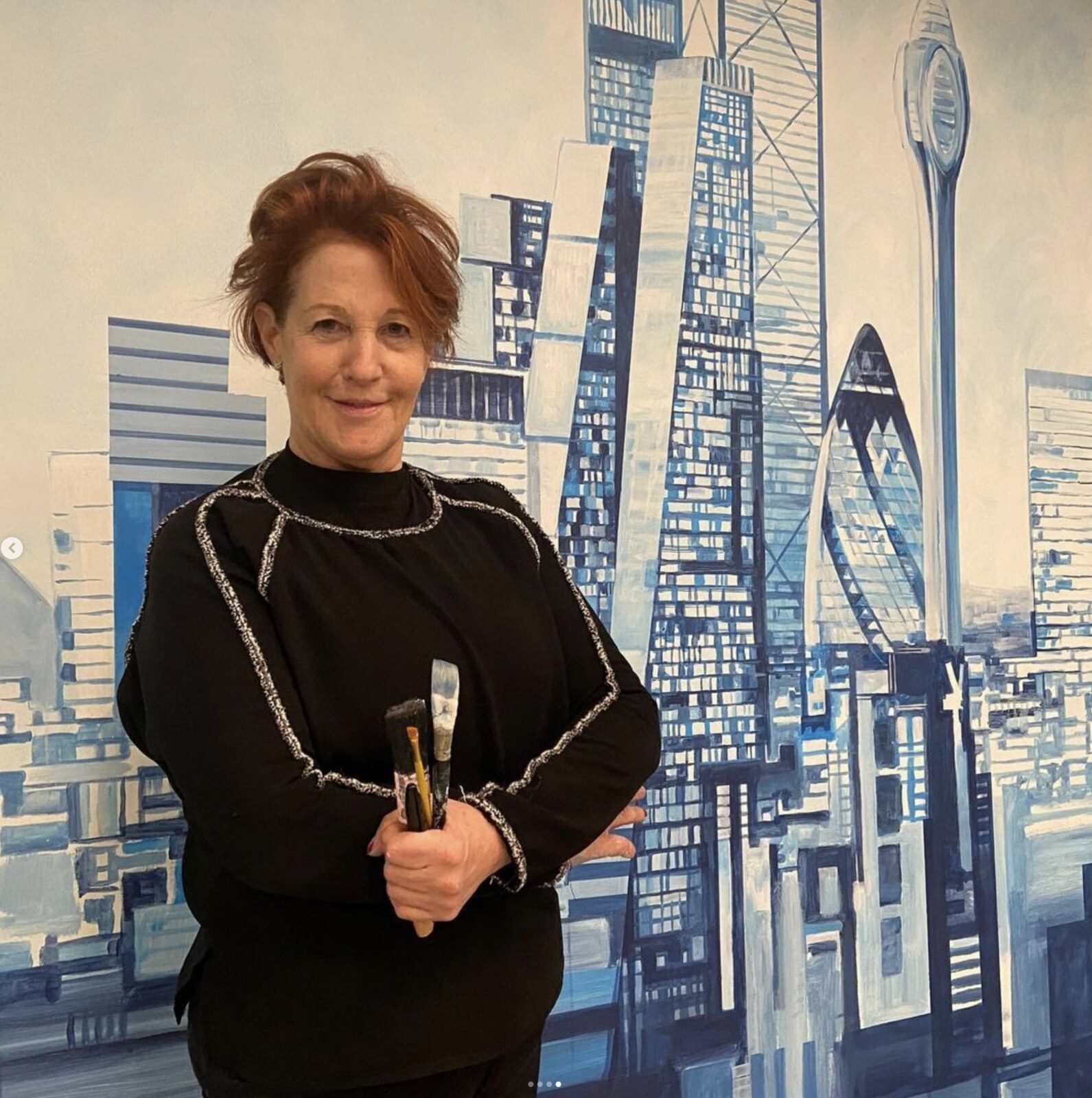Louise Garman holding paintbrushes in front of a large blue cityscape mural
