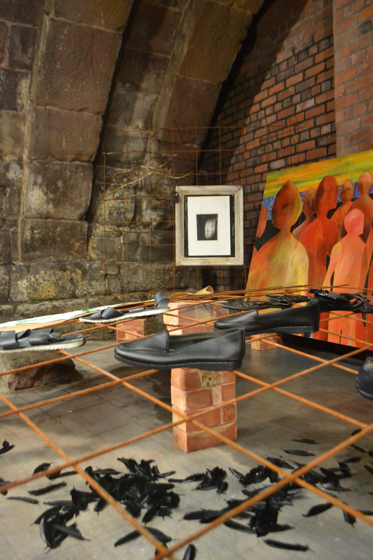 Shoes suspended on a rebar grid with black feathers scattered on the floor, inside the sandstone undercroft at Hanging Bridge, Manchester Cathedral