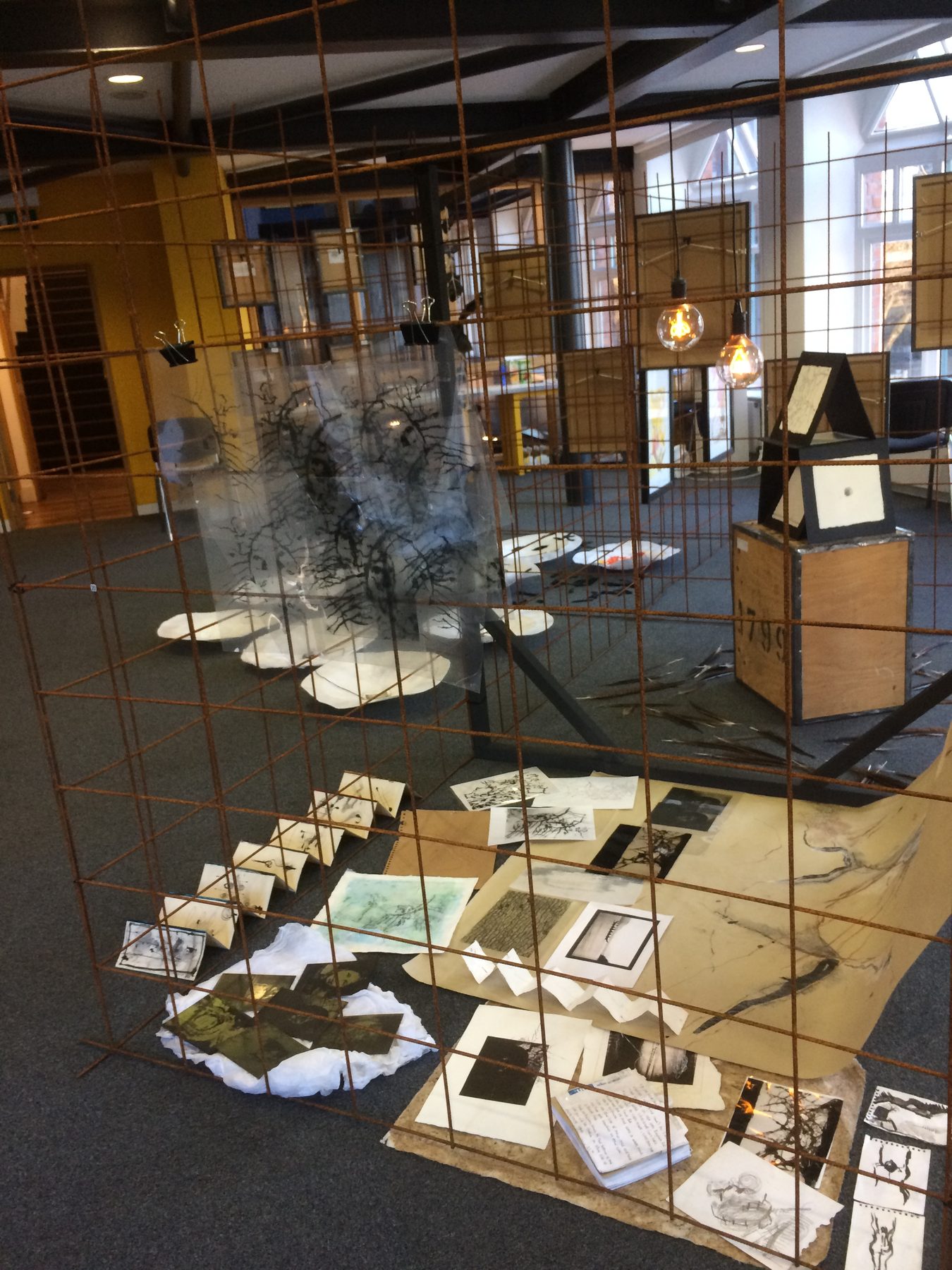 A rusted wire grid filled with framed charcoal tree drawings and a floor spread of photographs and sketches, exhibited at Manchester Cathedral