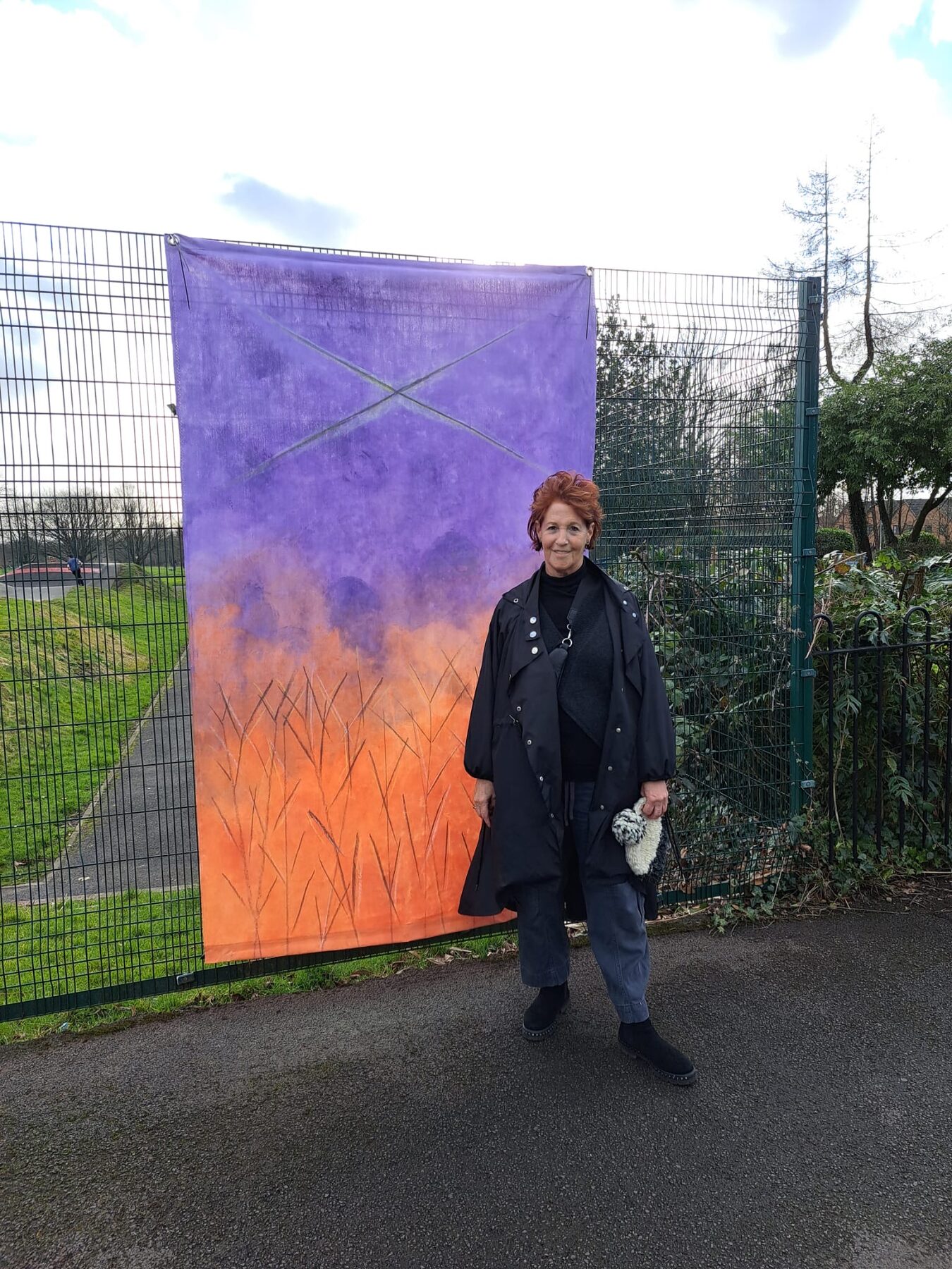Louise Garman beside one of her Human Nature fabric hangings, a purple and orange landscape, on the fence at Peel Park