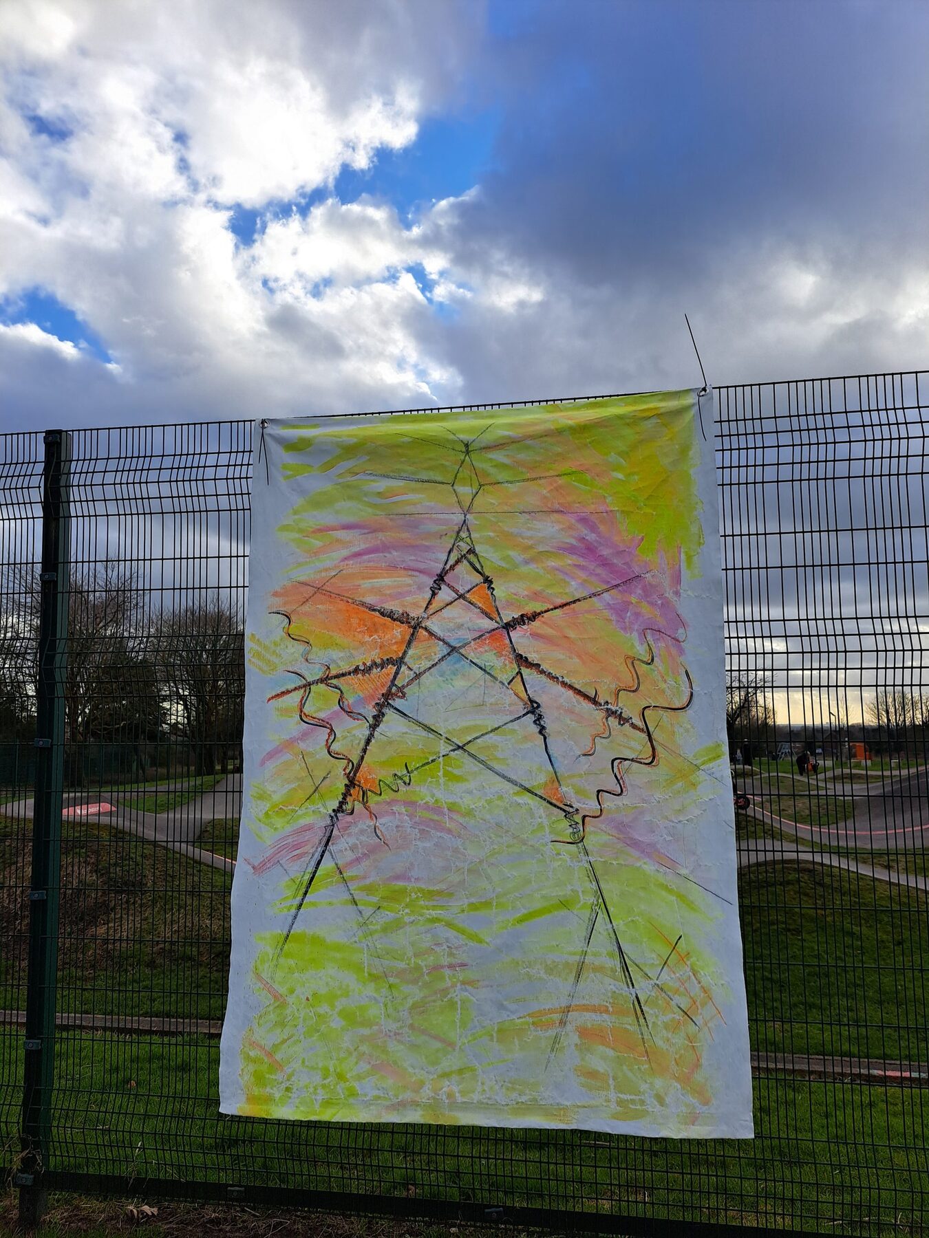 A green, pink and orange fabric hanging with a star-shaped linear design, displayed on the perimeter fence at Peel Park