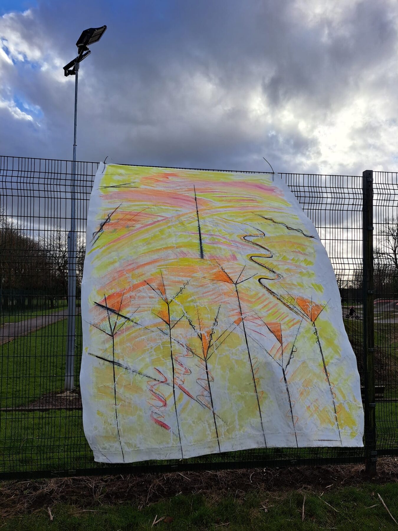A yellow, orange and pink fabric hanging with tall tree-like forms, displayed on the perimeter fence at Peel Park