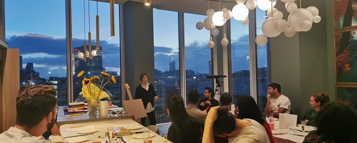 Louise Garman leading a watercolour workshop at dusk, with students painting at a table overlooking the Manchester skyline