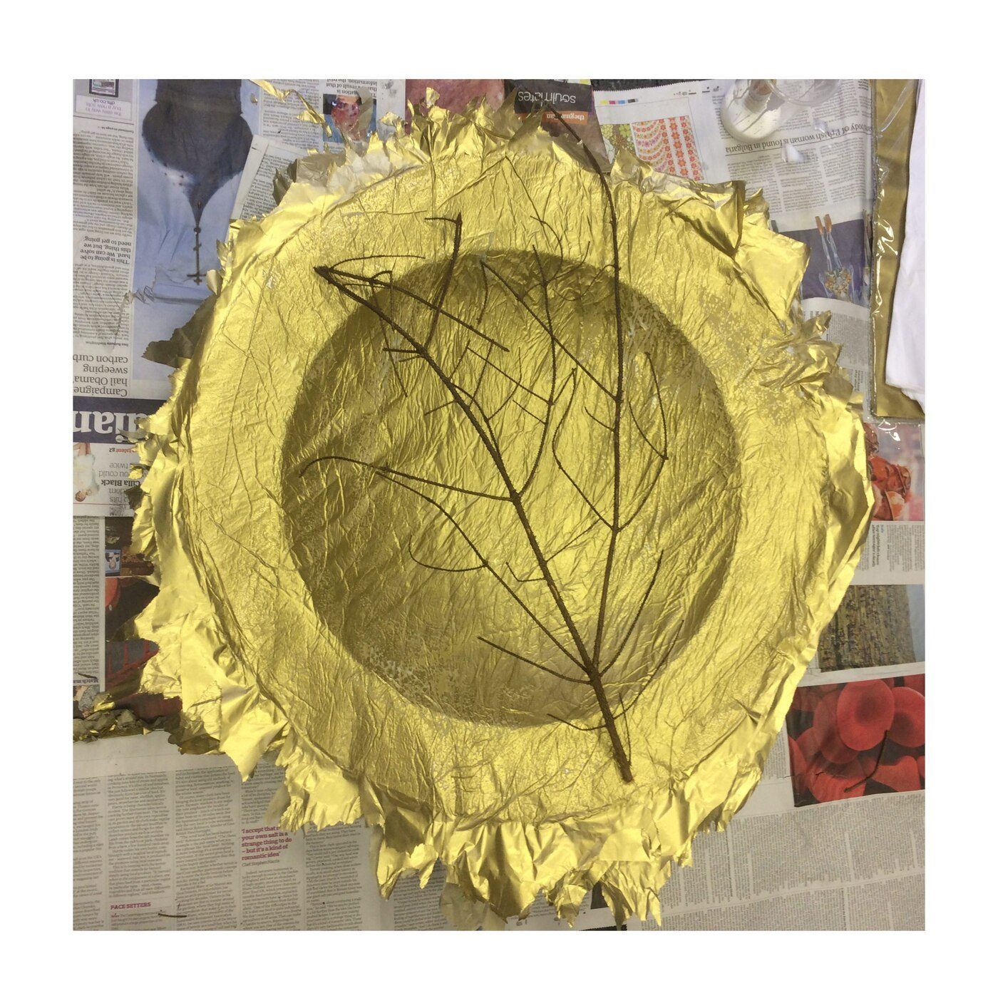 Gold foil circular sculpture with branch, on a newspaper ground