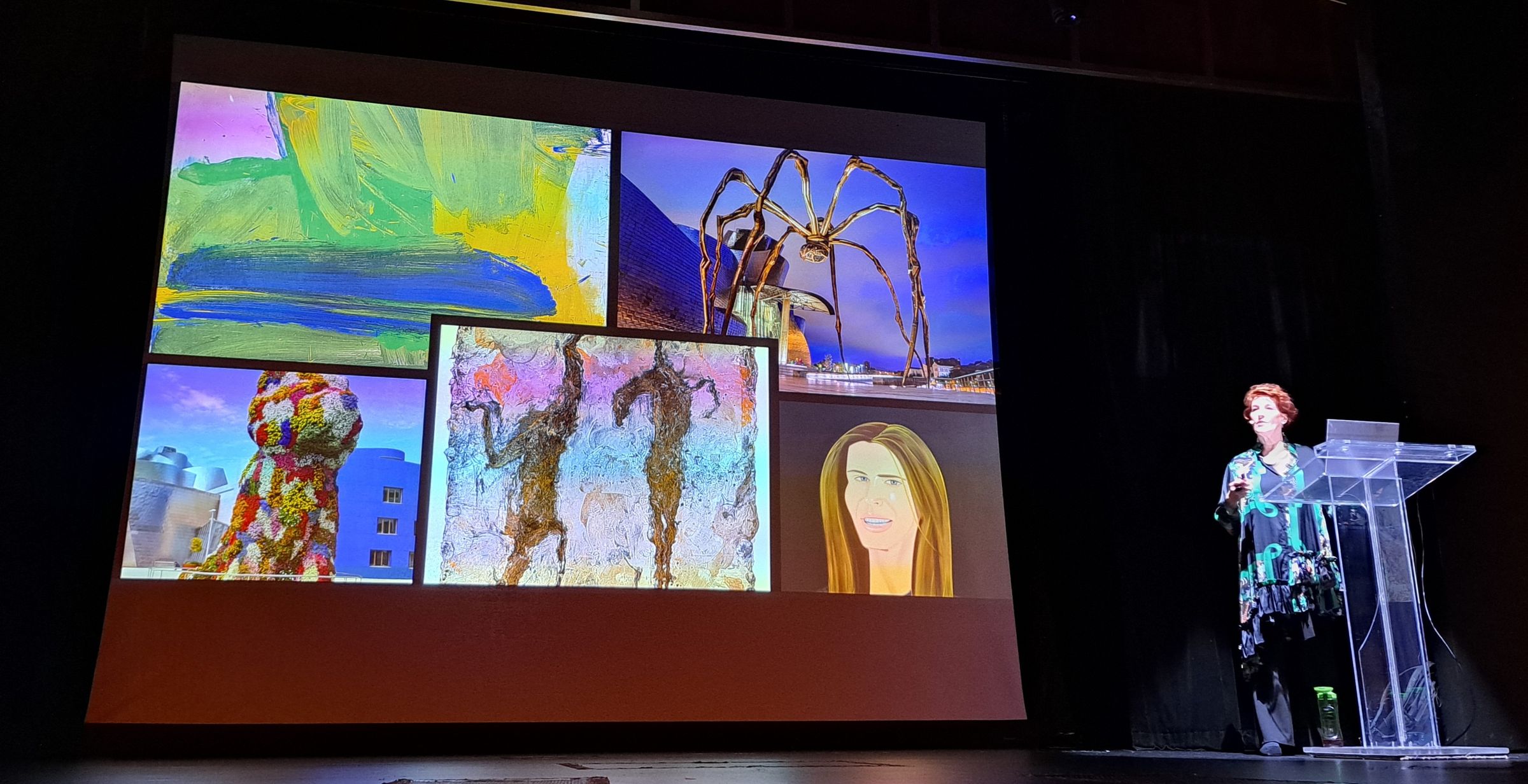 Louise Garman speaking at a podium beside a projected slide collage including Louise Bourgeois's spider sculpture at the Guggenheim Bilbao, Jeff Koons's Puppy, cave art figures and an abstract painting